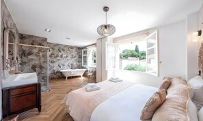 A bedroom with a large bed, a vanity with a mirror, a bathtub, and a window with a view of greenery. - POOL FRENCH VILLA WITH GYM , SAUNA - IN THE HEART OF CANNES- 250 SQM (Cannes)