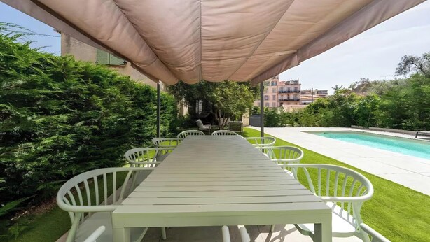 Un área de estar al aire libre con una mesa y sillas bajo un toldo, una piscina y vegetación.