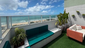 A balcony with a view of the beach, a green sofa, and potted plants. - Pertinho do Mar, Brisa da Praia e 2 Quartos (Fortaleza)