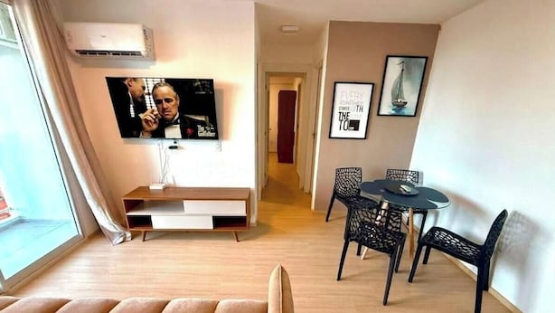 A modern living room with a flat-screen TV mounted on the wall, a dining area with a round table and chairs, and a hallway leading to another room. - Pertinho do Mar, Brisa da Praia e 2 Quartos (Fortaleza)