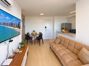 A modern living room with a sofa, a dining area with chairs, a kitchen with a refrigerator and microwave, and a TV displaying a beach view. - Pertinho do Mar, Brisa da Praia e 2 Quartos (Fortaleza)