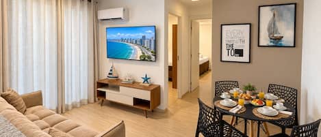 Una sala de estar moderna con un sofá, una mesa de comedor dispuesta para el desayuno y un televisor que muestra un paisaje urbano.