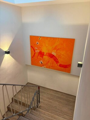 A staircase with a wooden floor and metal railings, leading to a room with a white wall and a large orange abstract painting. - Eibisch Apartments (Passau)