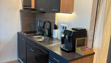 A compact kitchen with a modern design, featuring a built-in oven, a coffee machine, and a small wooden cabinet.