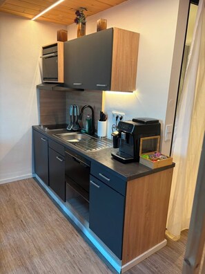 Una cocina compacta de diseño moderno, con horno integrado, cafetera y un pequeño armario de madera.