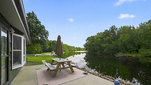 Järvenrantaterassi, jossa on piknikpöytä ja aurinkovarjo, ja sen ympärillä rehevää kasvillisuutta sekä tyyni järvi.