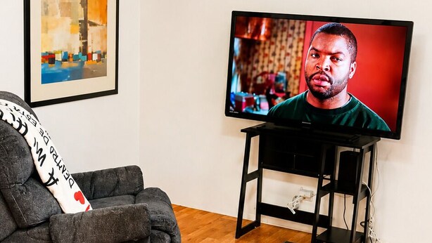 Ein Wohnzimmer mit Fernseher, einem grauen Sofa, einem schwarzen Teppich und einem Deckenventilator.