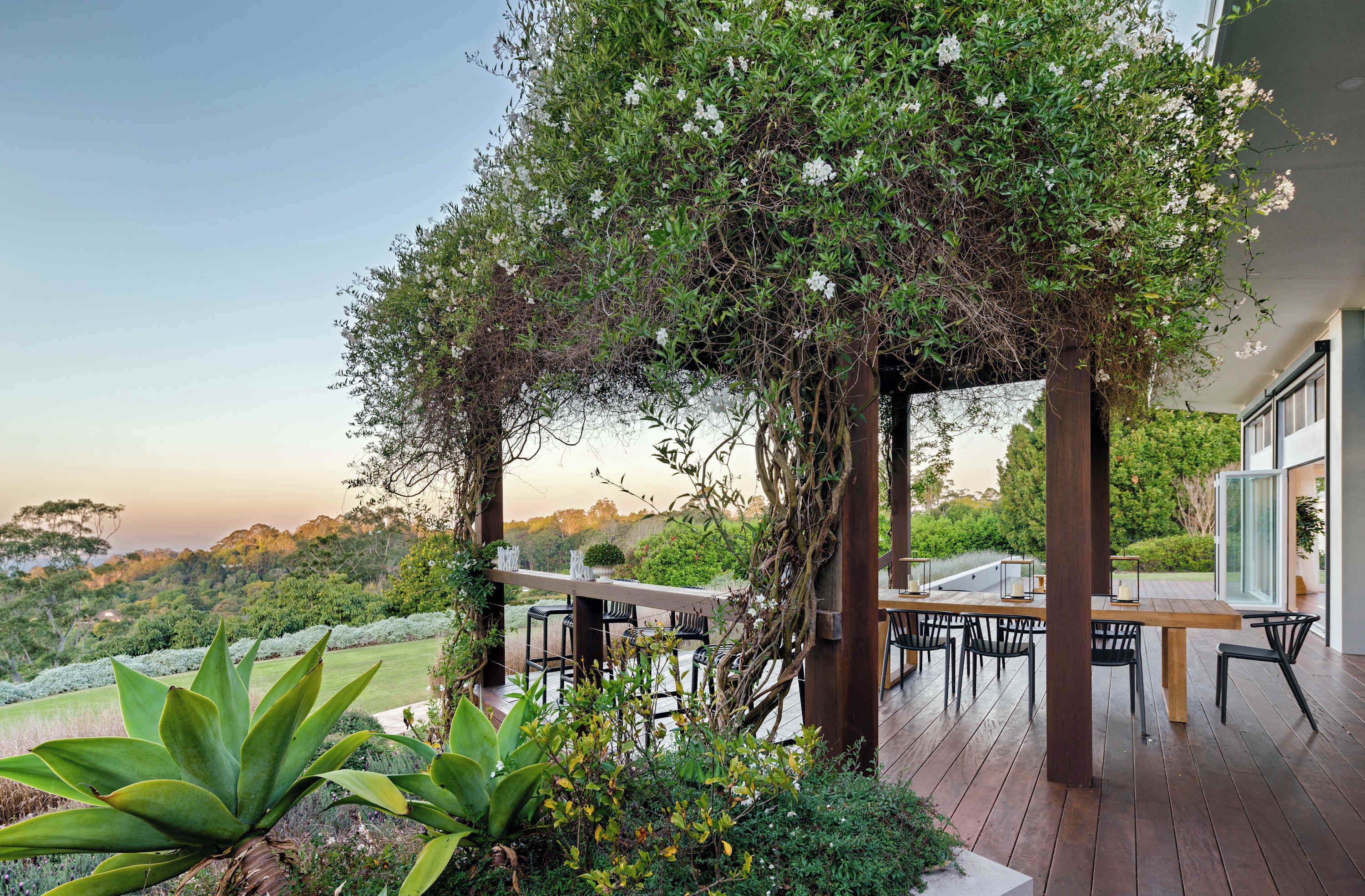 An outdoor patio with a wooden table and chairs, surrounded by greenery and a view of the landscape.