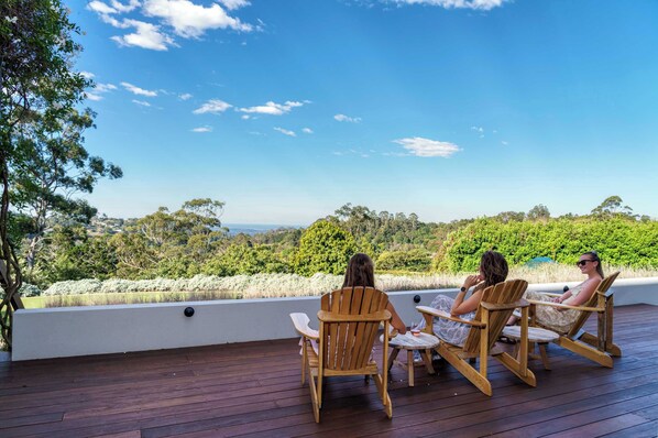 A wooden deck with two wooden chairs and a small table, overlooking a scenic landscape with trees and a clear sky.