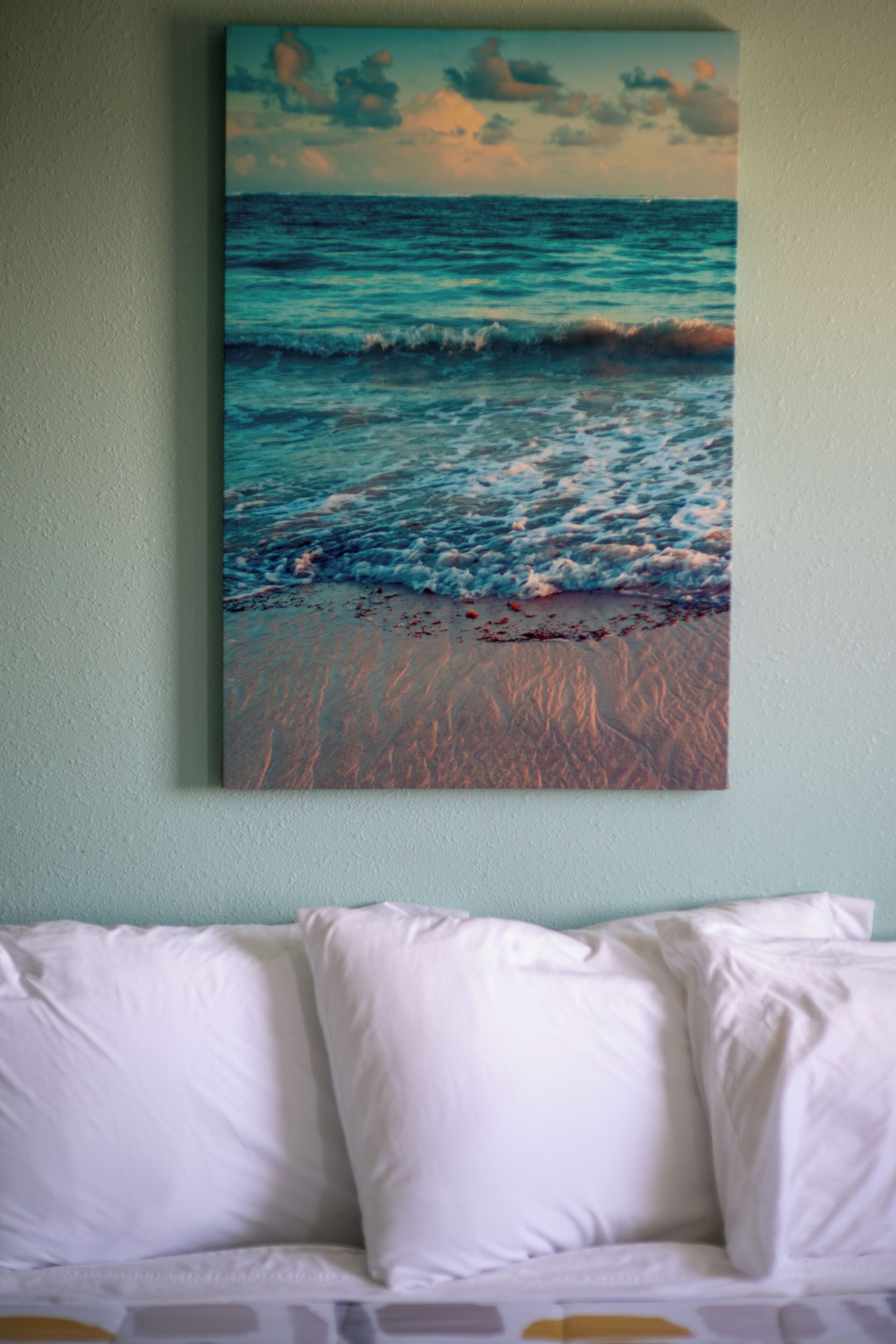 A painting of a beach scene with waves and a sunset on a wall above a bed with white pillows.