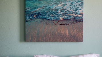 Pintura de una escena de playa con olas y un atardecer sobre una pared, encima de una cama con almohadas blancas.