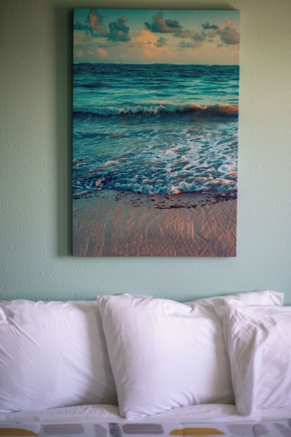 A painting of a beach scene with waves and a sunset on a wall above a bed with white pillows.