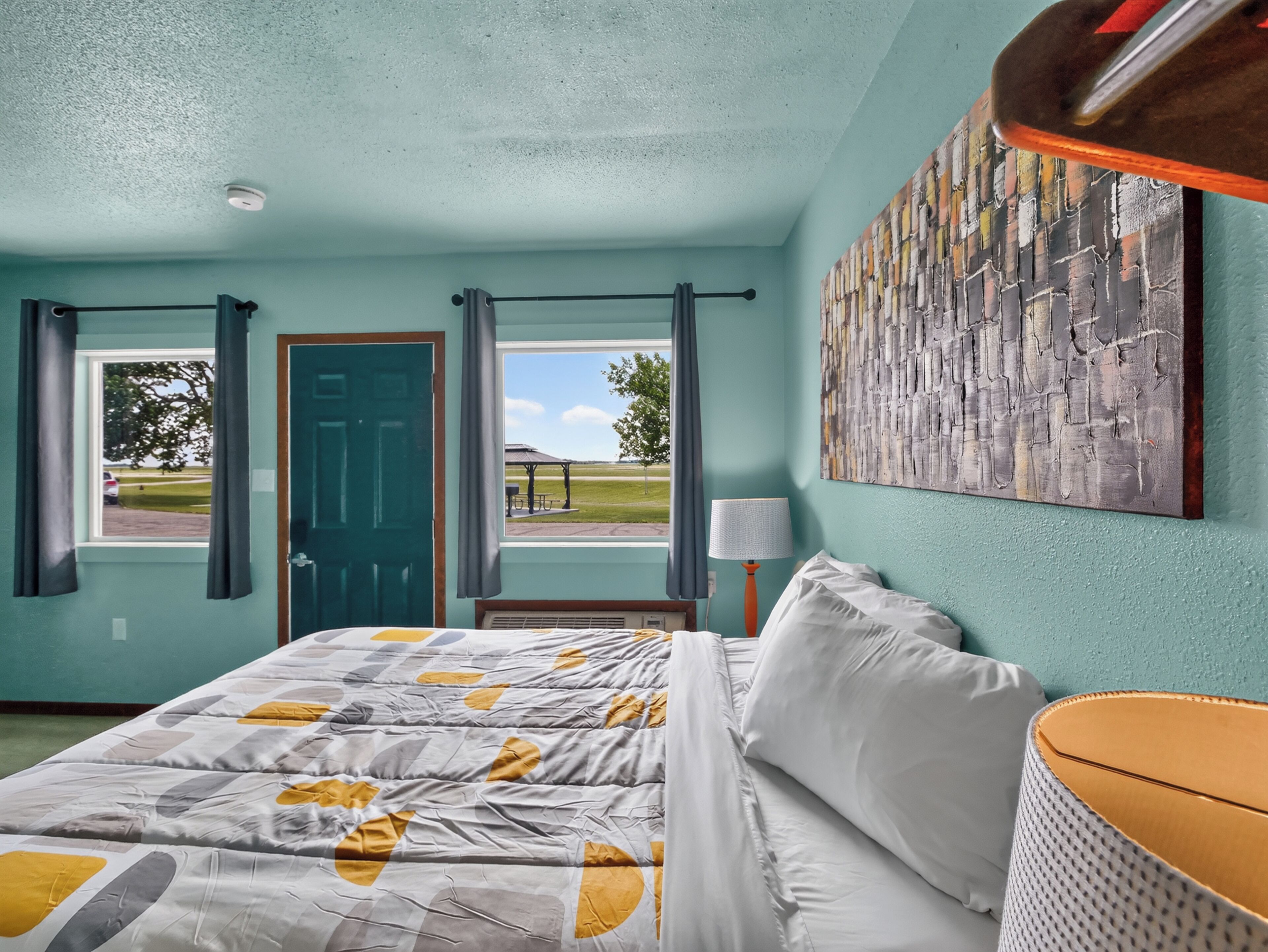 Una habitación con cama, ventana con cortinas, puerta, lámpara de pared y un cuadro.