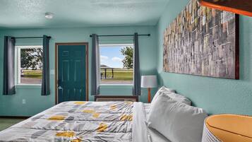 Una habitación con cama, ventana con cortinas, puerta, lámpara de pared y un cuadro.