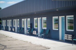 Un edificio con una fila de ventanas y puertas, mesas y sillas al aire libre, y un cielo despejado.
