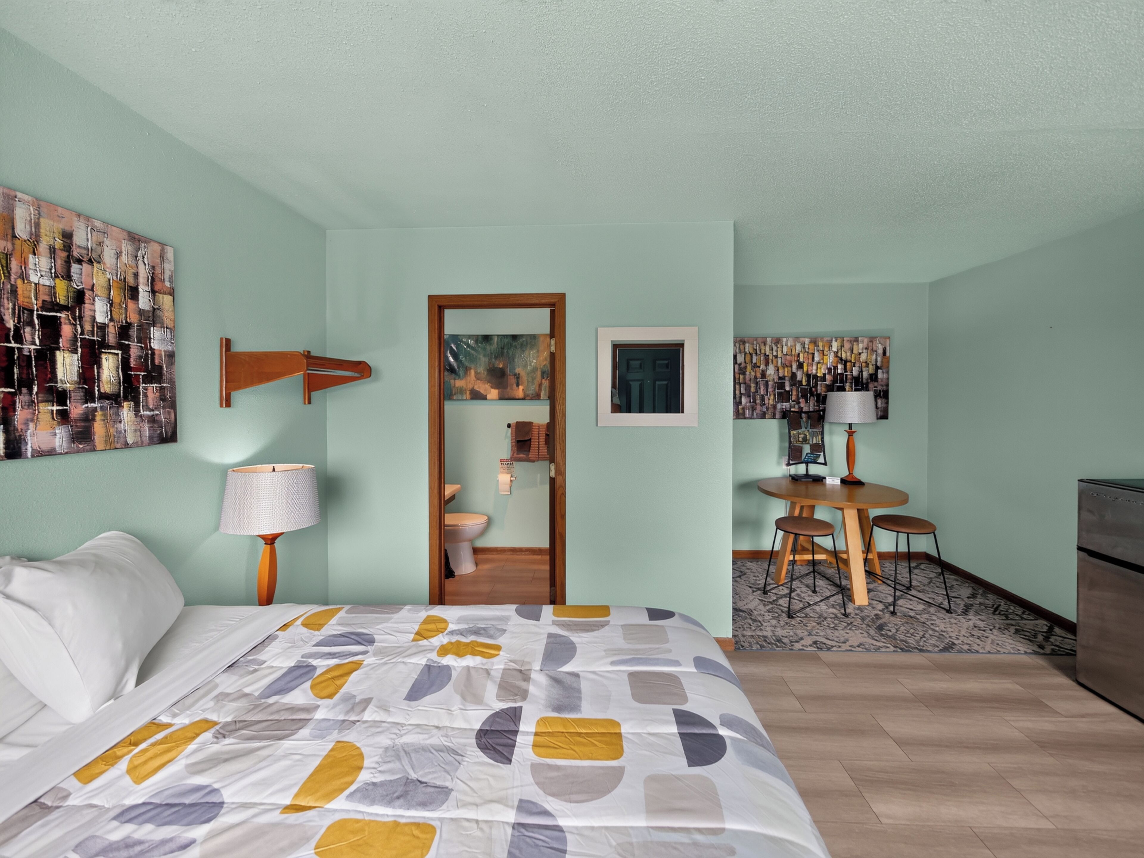 Una camera d'albergo con un letto, una scrivania, una sedia, una lampada e opere d'arte alle pareti.
