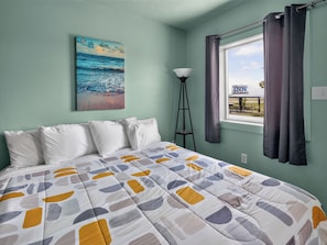 Un dormitorio con una cama, un cuadro de una escena de playa, una lámpara y una ventana con cortinas.