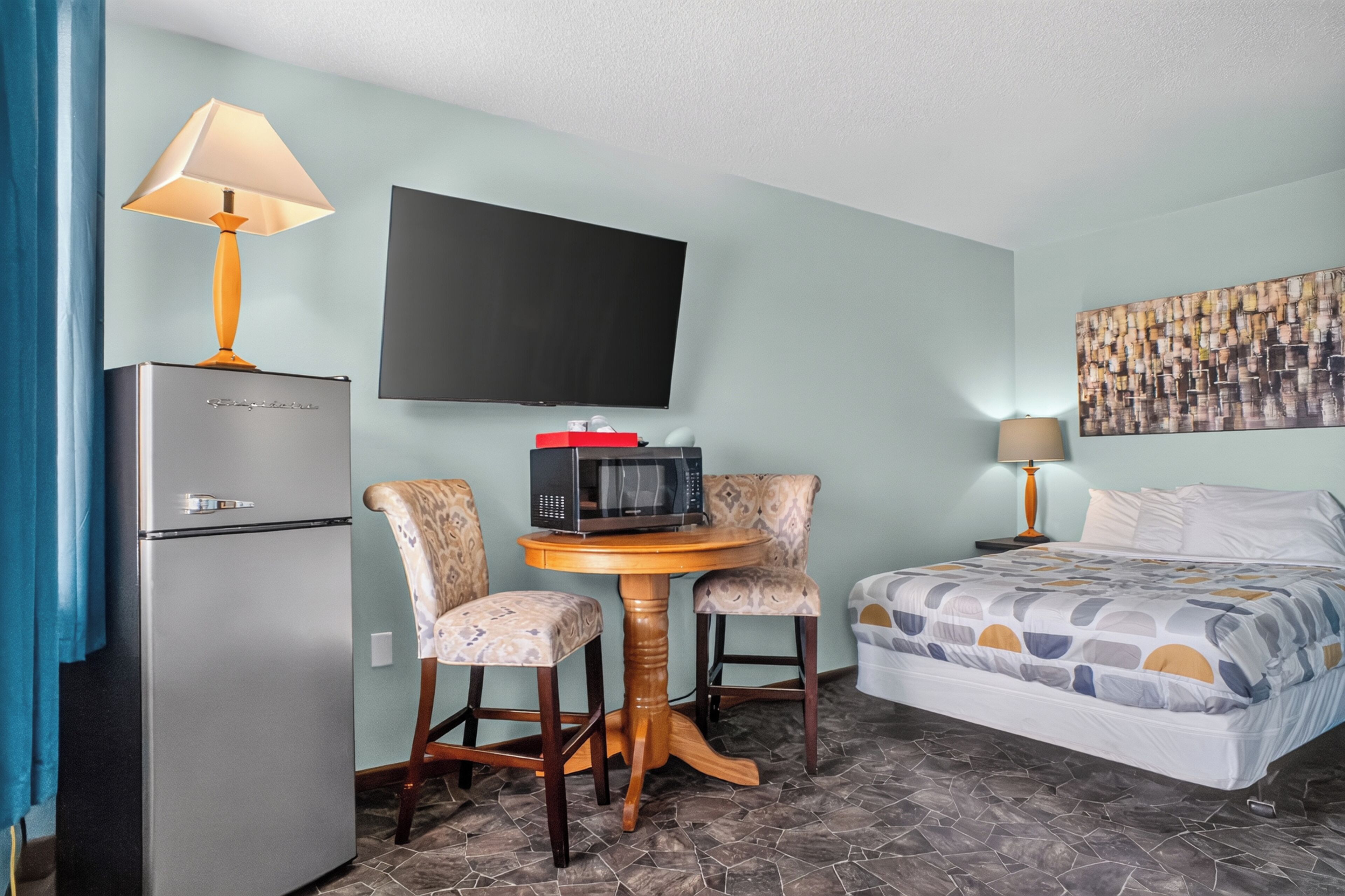 A hotel room with a bed, a television, a refrigerator, a table with two chairs, and a wall-mounted artwork.