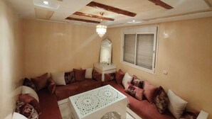 A living room with a sofa set, a coffee table, a chandelier, and a window with shutters. - Rachid (Agadir)
