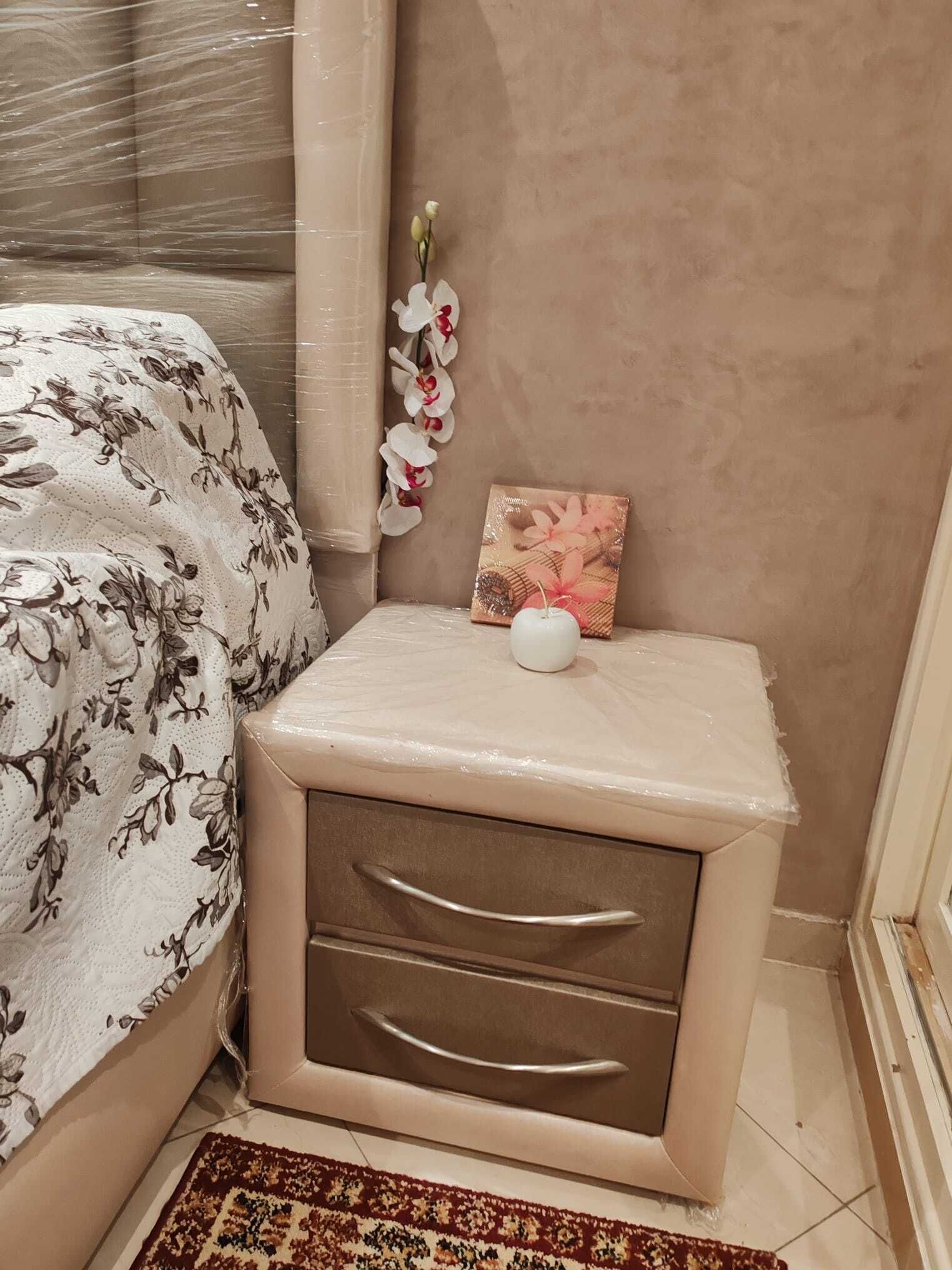 A bedroom with a bed, a nightstand with drawers, a small table with a picture, and a decorative orchid plant.