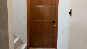 Una porta de fusta amb un ull de claus i un pany, situada en una paret senzilla amb una presa elèctrica visible.