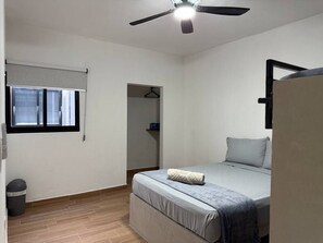 Een slaapkamer met een bed, een plafondventilator, een raam met jaloezieën, een deur en een afvalbak.