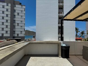 Een dakterras met een betonnen vloer, een zwarte prullenbak en uitzicht op een gebouw en de zee.