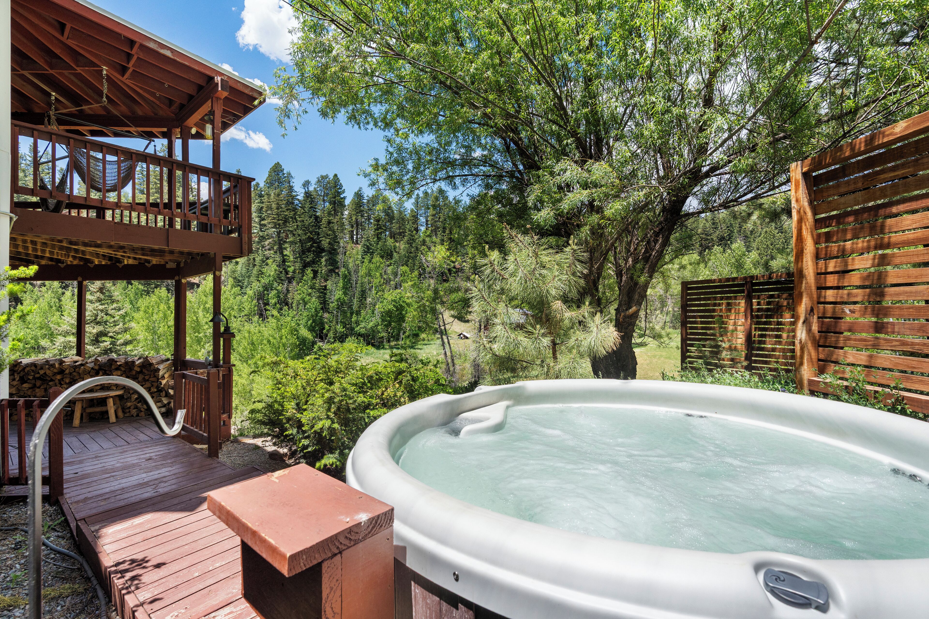 Un jacuzzi en una terraza de madera con un pérgola de madera y vista a un bosque.