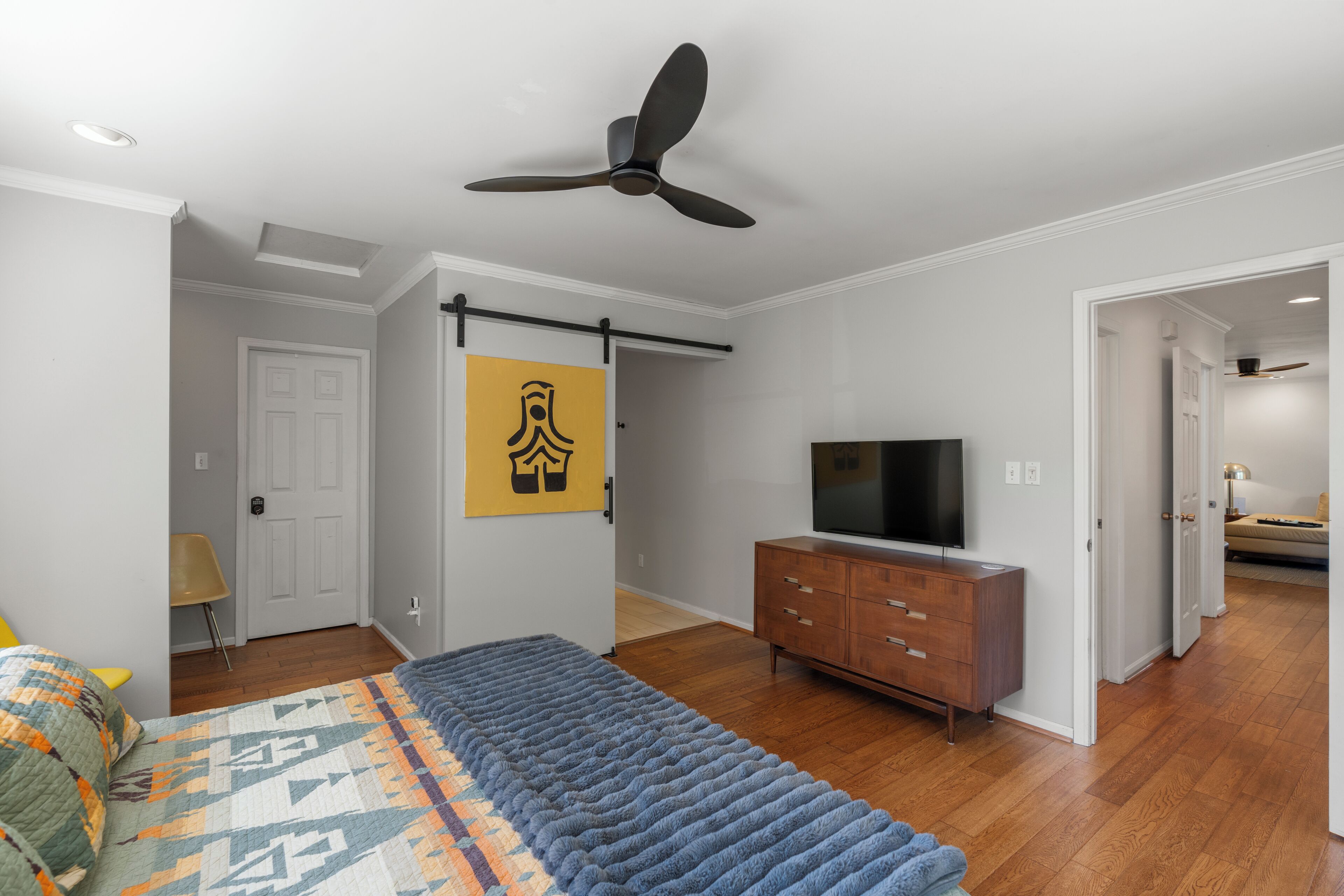 A bedroom with a bed, a TV, a ceiling fan, a dresser, and a door leading to another room.