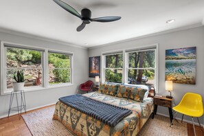 Ein Schlafzimmer mit einem Bett, einem roten Stuhl, einem gelben Stuhl, einem Deckenventilator und großen Fenstern mit Blick auf Grünflächen.