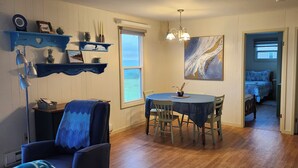 A room with a dining table, chairs, a blue chair with a blue cover, a painting on the wall, and a bed in the adjacent room.