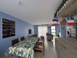 A room with a dining table, chairs, a sofa, a TV, and a kitchen area with wine bottles. - Maison climatisée avec jardin (BISCARROSSE)