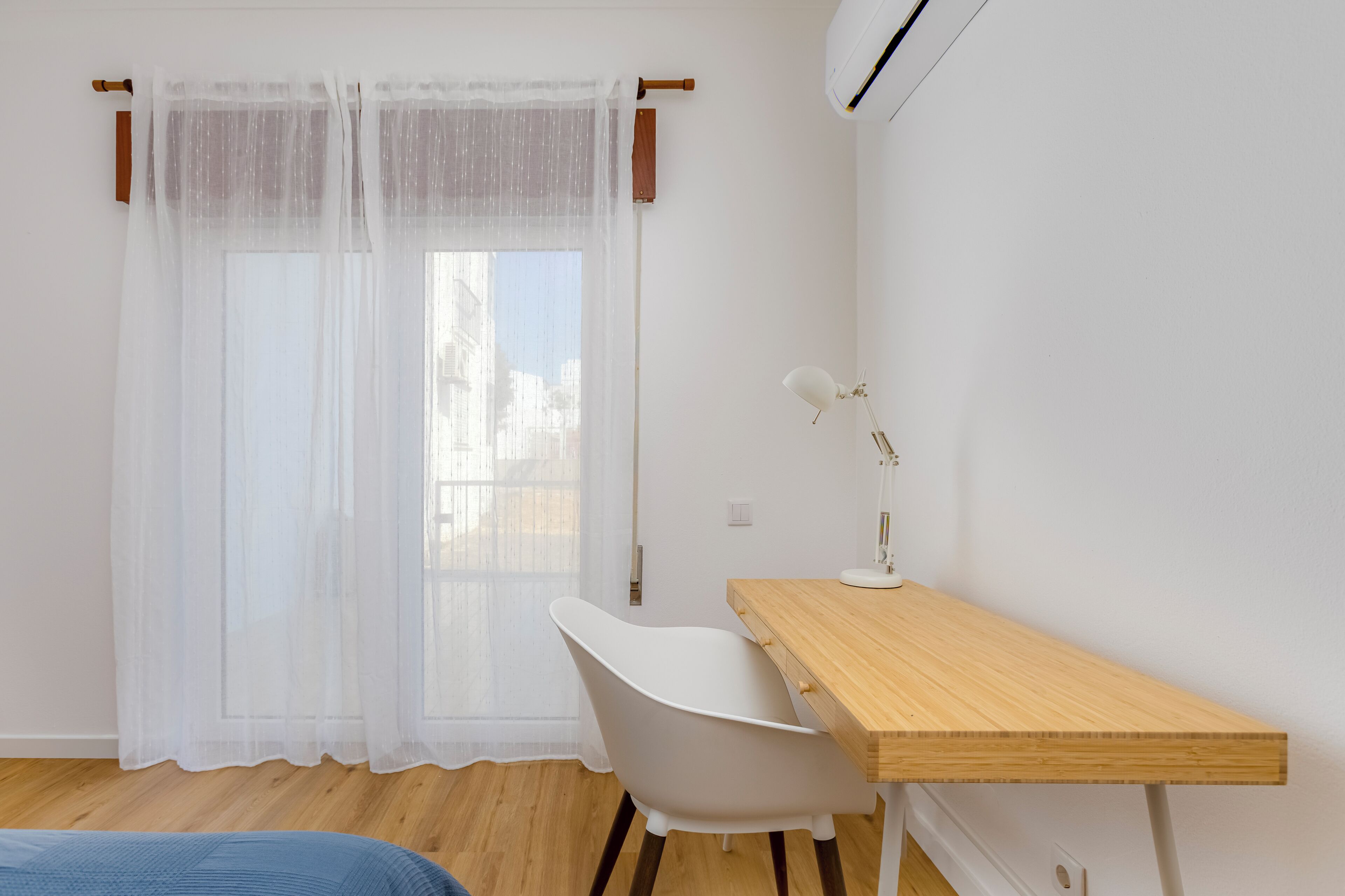 Een kamer met een houten bureau, een witte stoel en een raam met dunne gordijnen.