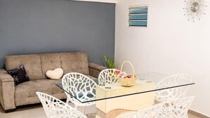 A modern living room with a glass-top dining table, white chairs, a ceiling fan, a sofa, and a wall clock. - Jasmin: Spacious, quiet apartment with a pool for 6 (Le Gosier)