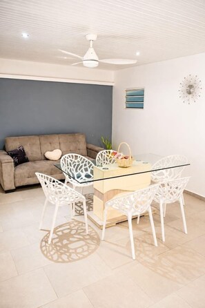 A modern living room with a glass-top dining table, white chairs, a ceiling fan, a sofa, and a wall clock. - Jasmin: Spacious, quiet apartment with a pool for 6 (Le Gosier)