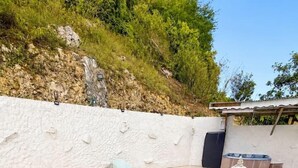 A rectangular swimming pool with a white tiled deck, surrounded by a white wall with a rocky hillside in the background. - Jasmin: Spacious, quiet apartment with a pool for 6 (Le Gosier)