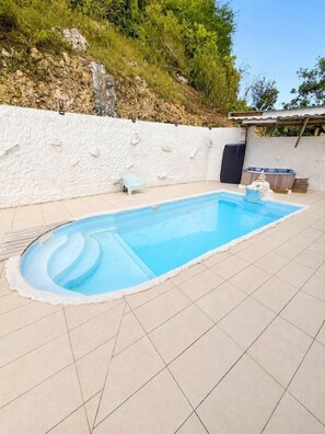 A rectangular swimming pool with a white tiled deck, surrounded by a white wall with a rocky hillside in the background. - Jasmin: Spacious, quiet apartment with a pool for 6 (Le Gosier)
