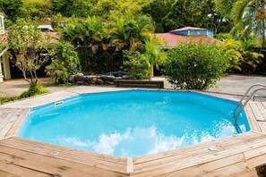 A rectangular swimming pool with a wooden deck surrounding it, set in a lush garden with various plants and trees. - Jasmin: Spacious, quiet apartment with a pool for 6 (Le Gosier)