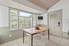Uma sala de jantar com uma mesa de madeira, cadeiras brancas, uma tigela, uma televisão fixada na parede e uma porta com janela.