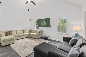 Una sala de estar con un sofá modular, un pequeño reposapiés, un tapete estampado, un ventilador de techo y un televisor montado en la pared que muestra 'ROYAL STAYS'.