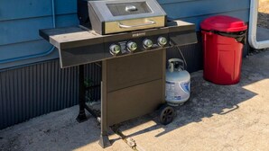 Una parrilla de gas portátil con panel de control y tanque de propano, sobre una superficie de concreto frente a una casa azul con una ventana.