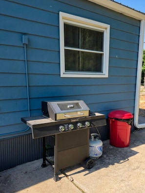 A portable gas grill with a control panel and a propane tank, placed on a concrete surface in front of a blue house with a window. - Enjoy the peaceful charm of this country villa with AC (Indianola)