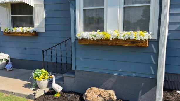 Una casa con fachada azul, un porche delantero de madera, una ventana con toldo blanco y macetas con flores en los alféizares.
