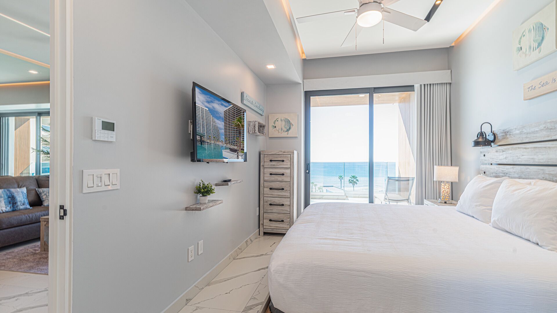 Quarto com uma cama grande, um ventilador de teto, uma TV na parede e vista para o mar pela janela.