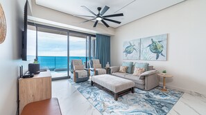 Un salon moderne doté d’une grande fenêtre, d’un ventilateur de plafond, d’un ensemble de canapés, d’une table basse et d’un tableau représentant des tortues de mer.