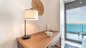 Camera d'albergo con una scrivania in legno, una lampada, un telecomando, una tazza e vista sul mare tramite una porta di vetro.