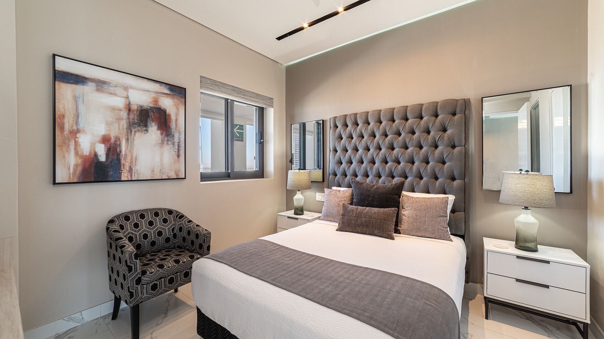 A modern bedroom with a tufted headboard, a large bed, a nightstand, a chair, and a painting on the wall.