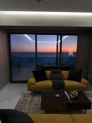 A modern living room with a yellow sofa, a coffee table, and a view of the ocean at sunset through large glass doors. - R1204 Oceanfront 3BR Condo with Stunning Views & Modern Comfort (Puerto Peñasco)