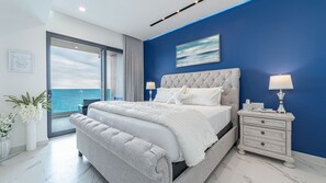 Une chambre avec un grand lit, une table de chevet, une fenêtre donnant sur la mer et un tableau encadré au mur.