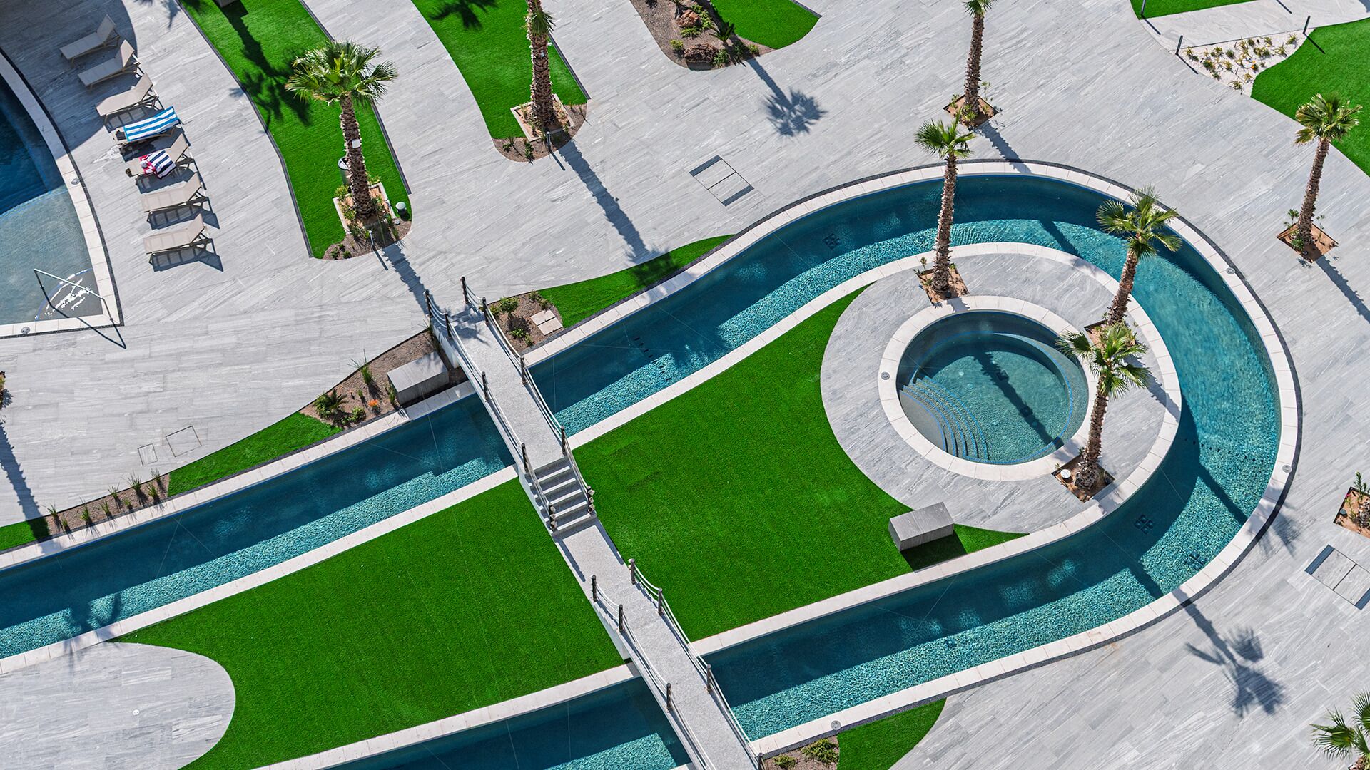 Vista aérea de una zona de piscina con una fuente sinuosa, rodeada de césped verde y palmeras.
